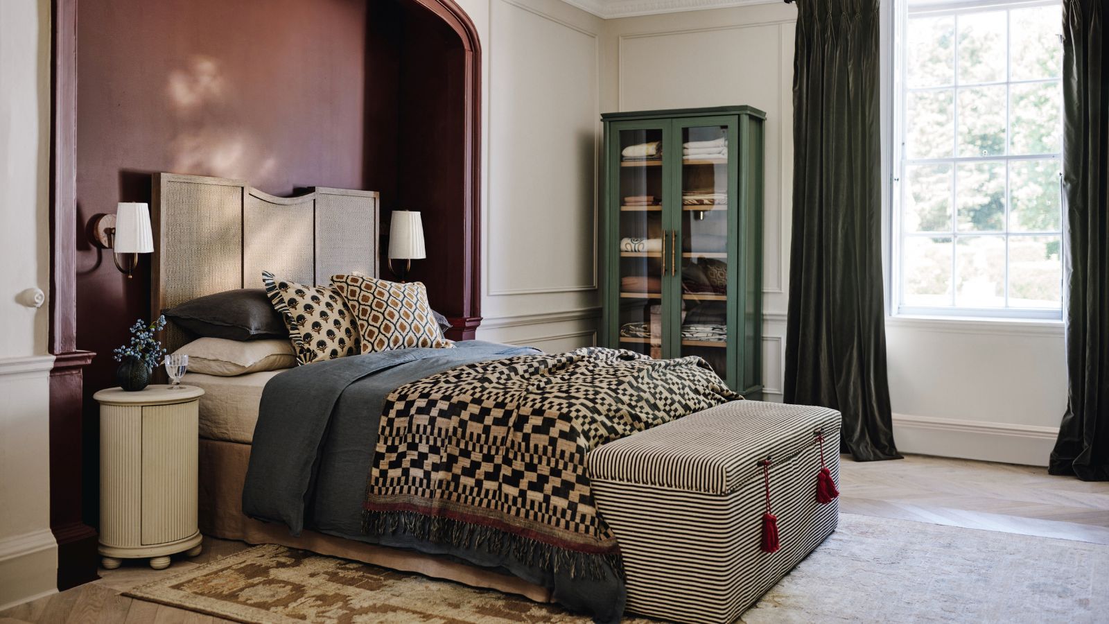 A wooden bed set into a red-painted alcove in a large bedroom. Patterned bedding on the bed and a grey pin-stripe ottoman at the foot.