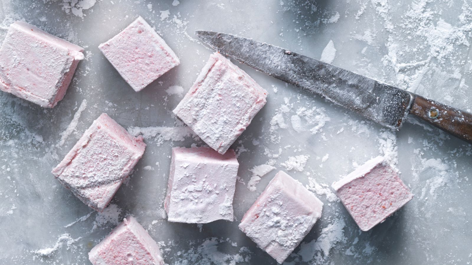 Cubes of pale pink cherry marshmallows on a cutting board with a knife