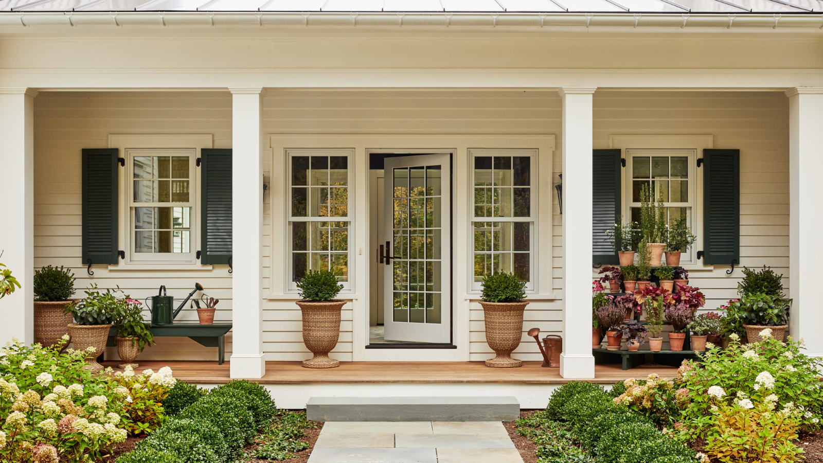 the exterior of a home with a large porch decorated with lots of plants and some plants in rattan planters