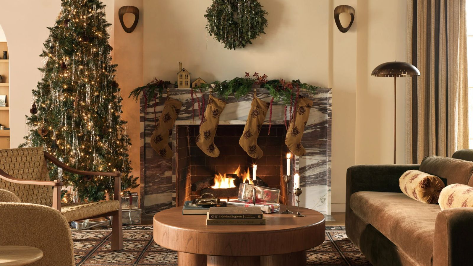 lulu and georgia christmas 2025 campaign imagery of a moody, warm living room with lit marble fireplace decorated with a garland, velvet stockings, a velvet couch, coffee table, green armchairs and a christmas tree