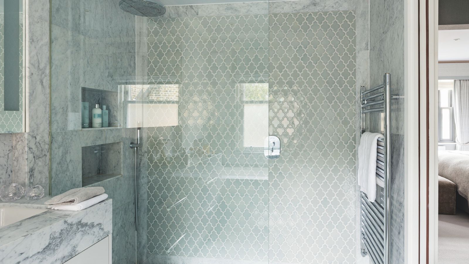 A marble and glass tiled shower with a shower niche, large glass screen, silver radiator and marble sink