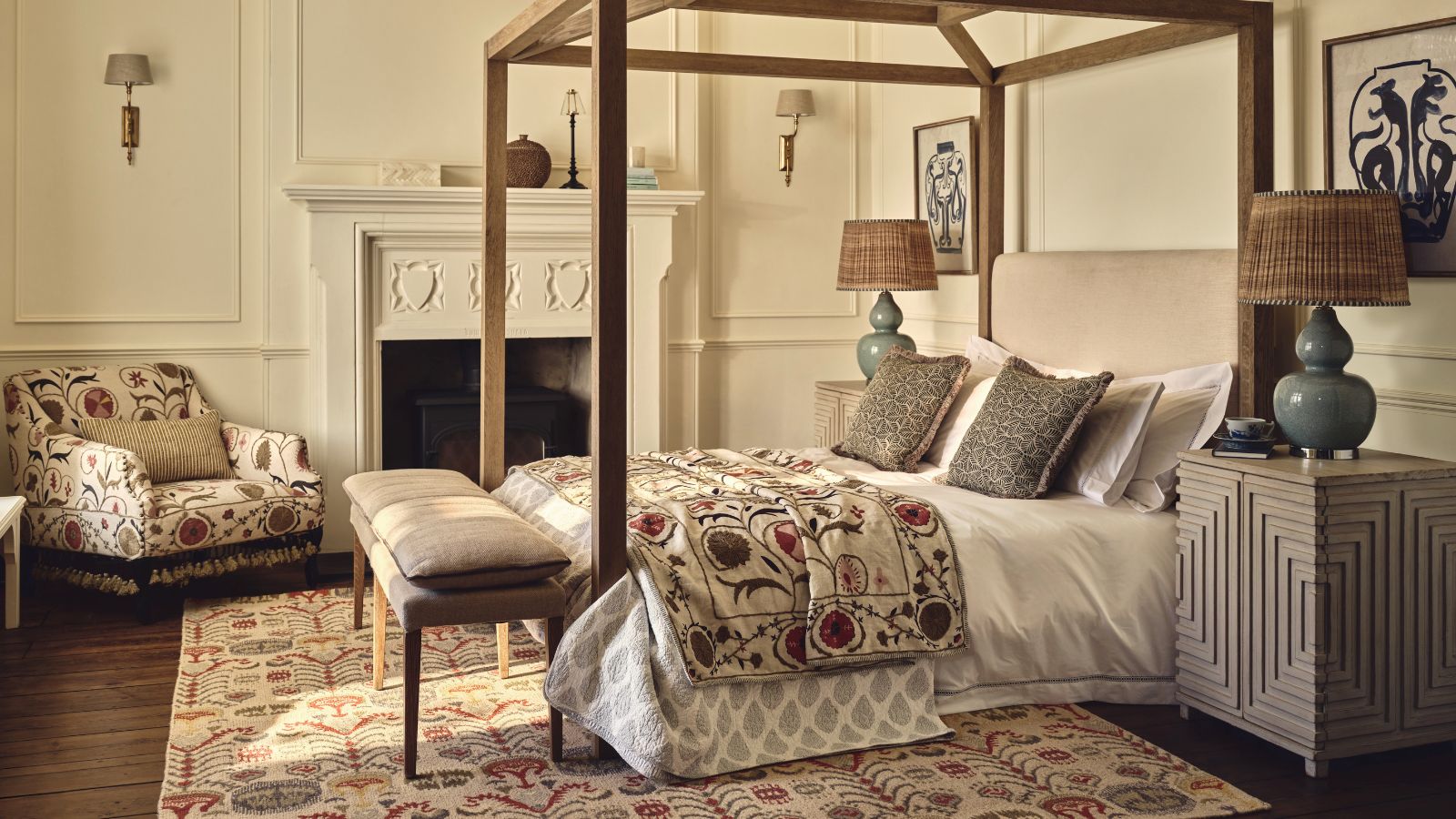 A traditional neutral bedroom with a wooden four poster bed, a bench at the base, and a chair in the corner of the room.