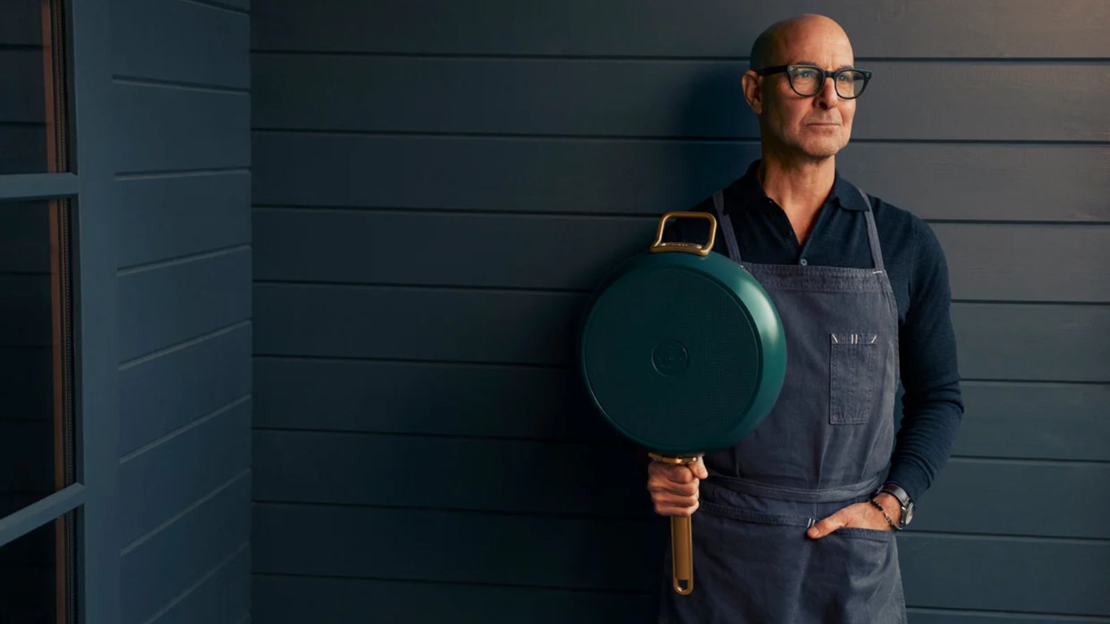 Stanley Tucci in blue apron holding a teal pan against a blue wood panelled backdrop