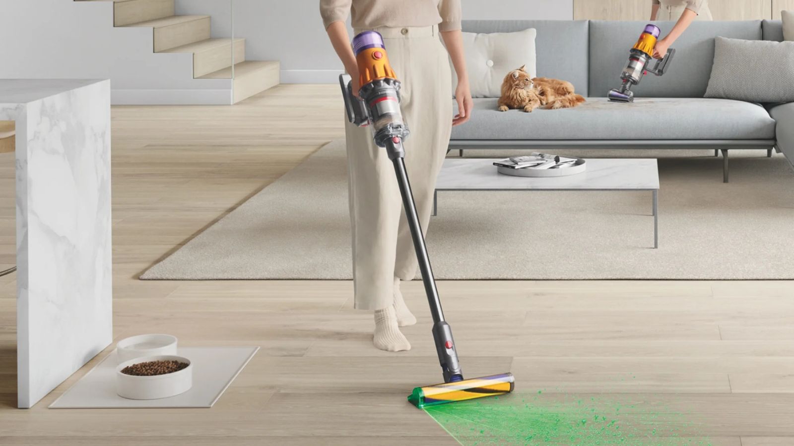 Person vacuuming hard floor with the Dyson V12 Detect Slim and green laser coming from floorhead. Somebody is pictured behind using the same vacuum in handheld mode next to a cat on a gray sofa, in a minimalist, neutral living room.