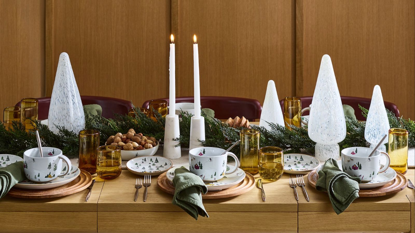 Christmas table setting on bare wooden table with glass trees, greenery, candles and coloured yellow glasses