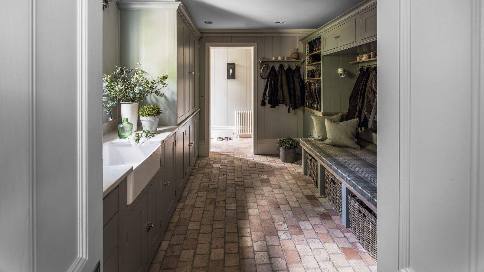 A tiled boot room with sage green cabinets, wicker baskets, a wooden bench with green cushions, a white sink, and green houseplants in white pots.