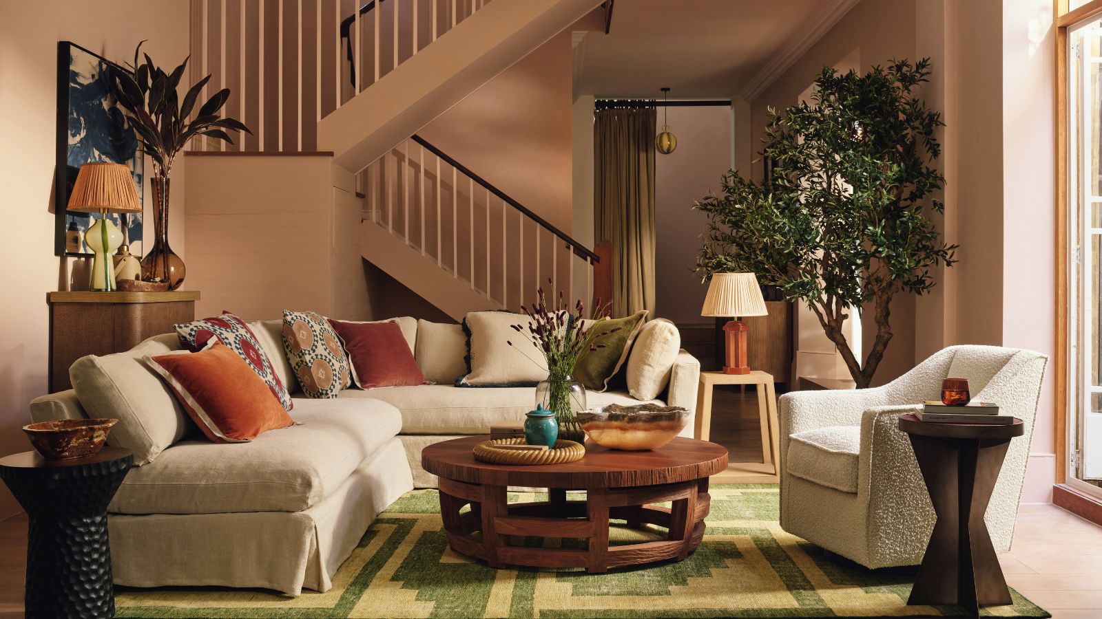 A warm-toned living room with a large cream L-shaped sofa, across from a matching armchair. A round wooden coffee table between them on a green and yellow rug.