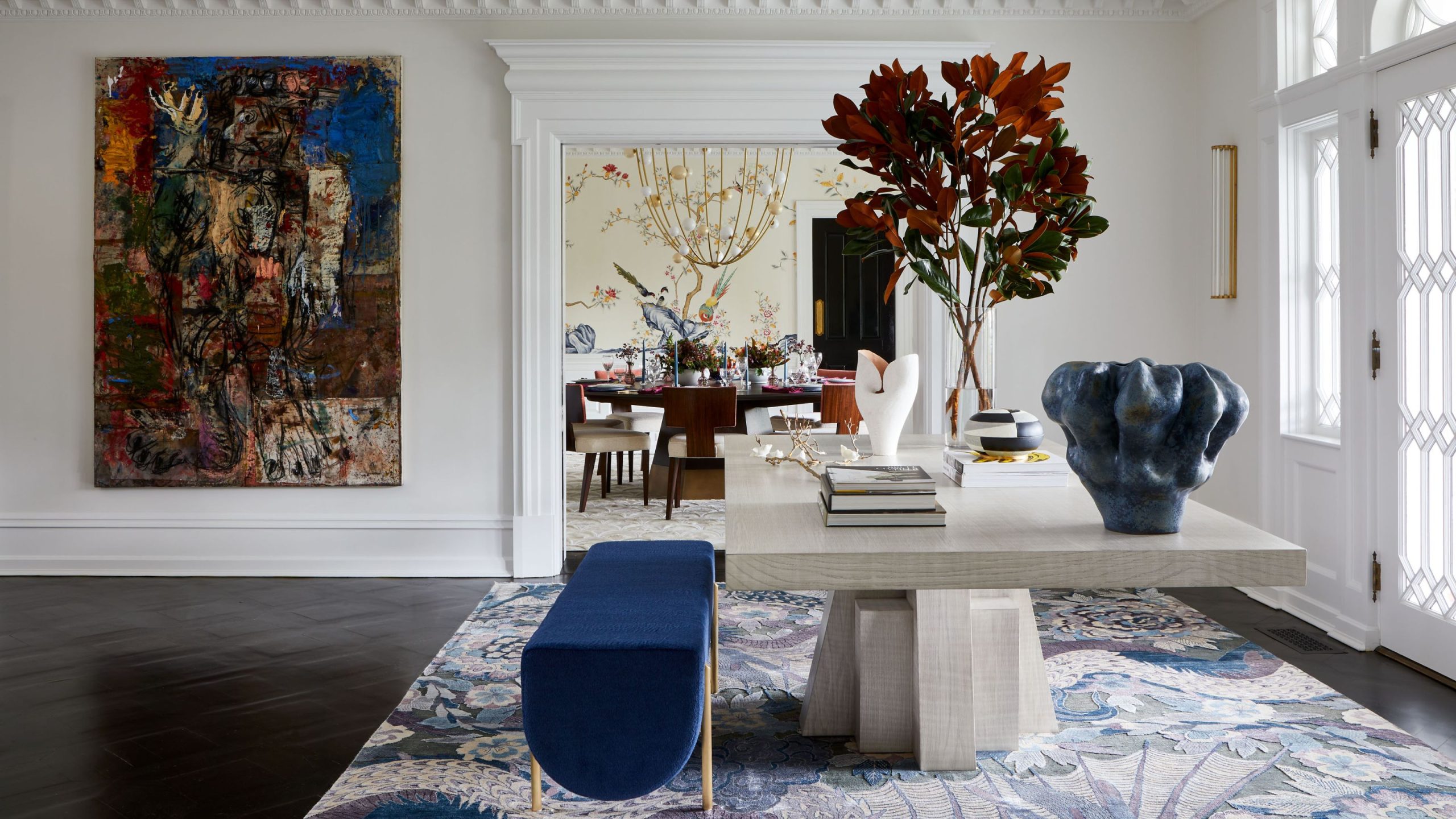Large entryway with white walls and a wooden floor. A stone table sits in the middle decorated with books and a large vase of foliage. There is a blue bench next to the table and the both sit on a patterned blue and white rug