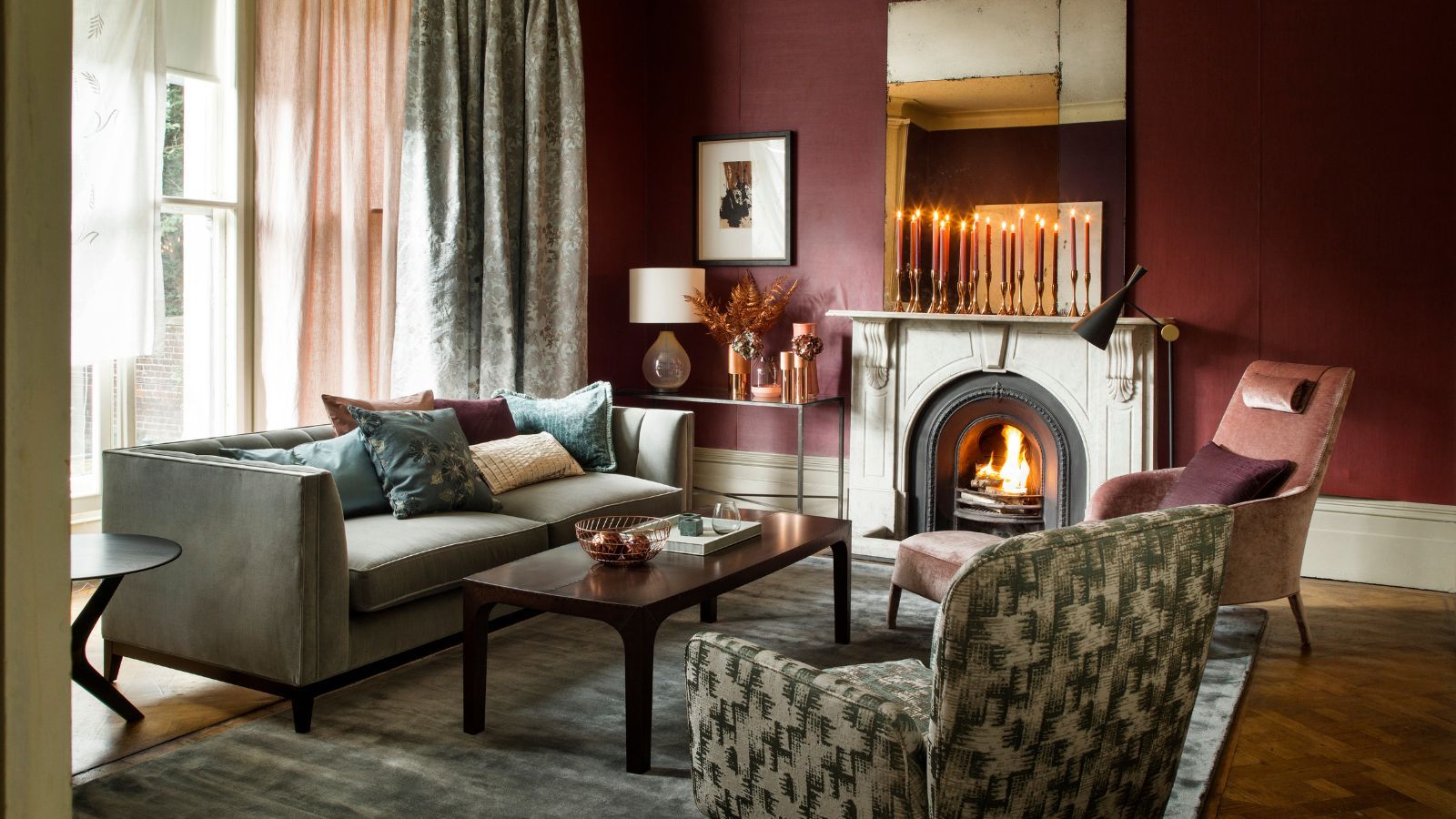 A cozy living room with burgundy walls, mismatched upholstered furniture, a large grey velvet rug, brown wooden coffee table, and fire burning in a stone fireplace with tall tapered candles burning on the mantelpiece above.