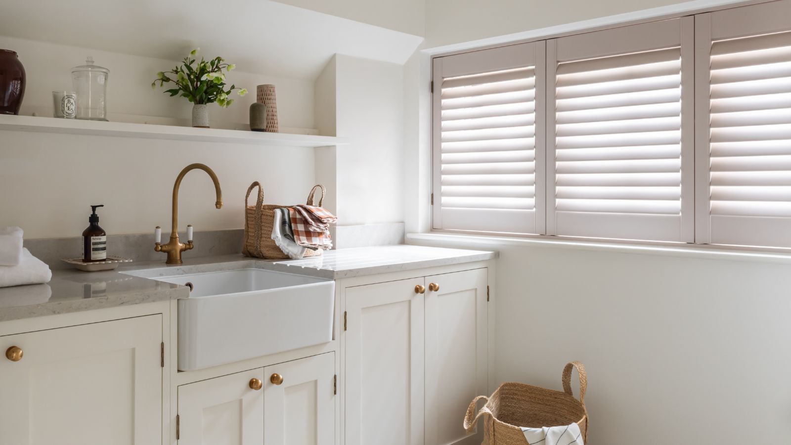 A white laundry room with white cabinets, walls, a sink, and shutters with gold doorknobs and faucets, and marble countertops.