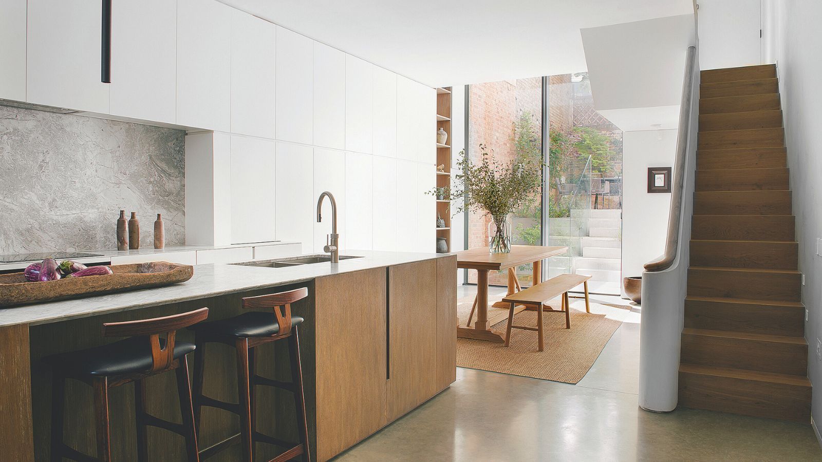 A white kitchen with a wooden kitchen island with in-built sink. A staircase on the right hand side leading up, and a wooden dining table and benches beside it. Large floor-to-ceiling glass at the back of the room showing a small patio.