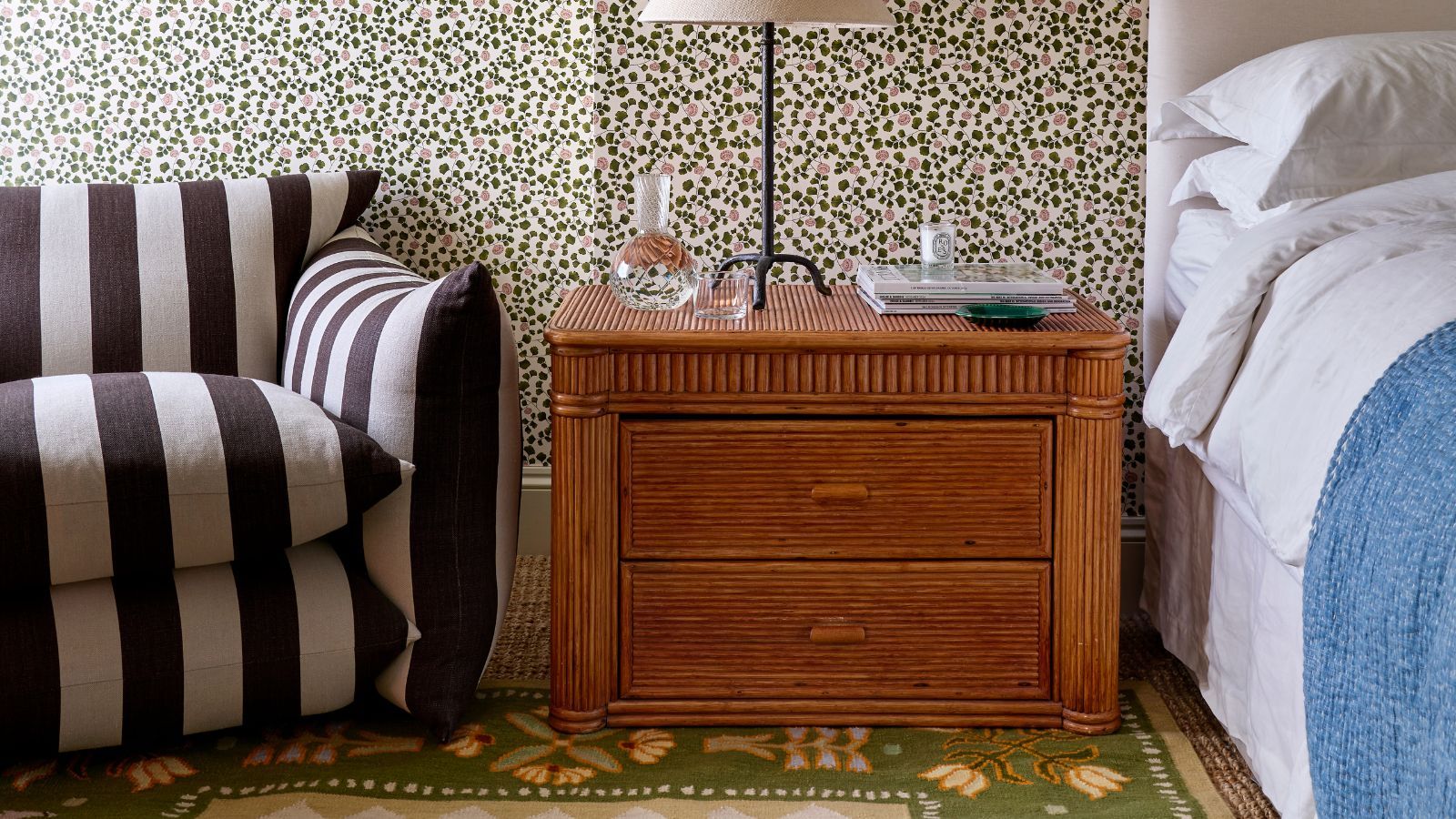 Lucy Williams Home x Pelican House rug collaboration showing a green floral flatweave rug in a floral wallpapered bedroom with white linens, a vintage bamboo nightstand and a black and white striped chair