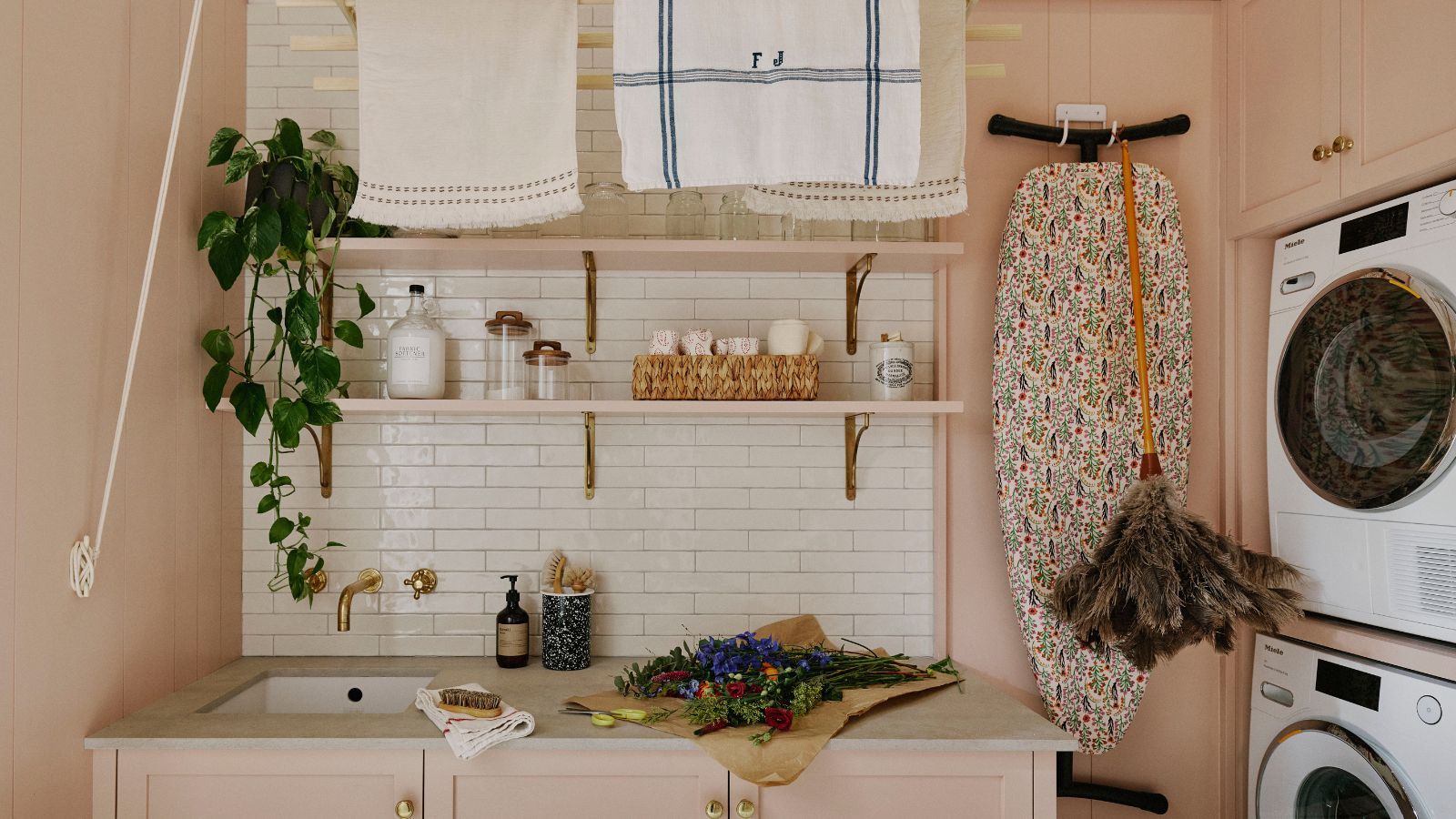 pink utility room with stacked machine and a sink area with pink cabinets and open shelves, white tiled backsplash, a hung ironing board and a laundry rack hung from the ceiling