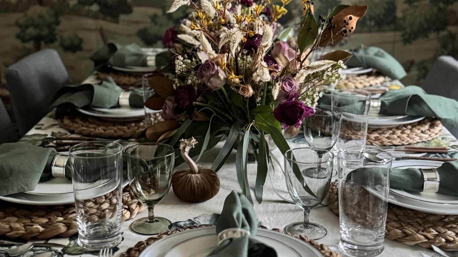 green themed table laid for thanksgiving with a white and green tablecloth, fall foliage centerpiece, wicker platemats and little velvet pumpkins