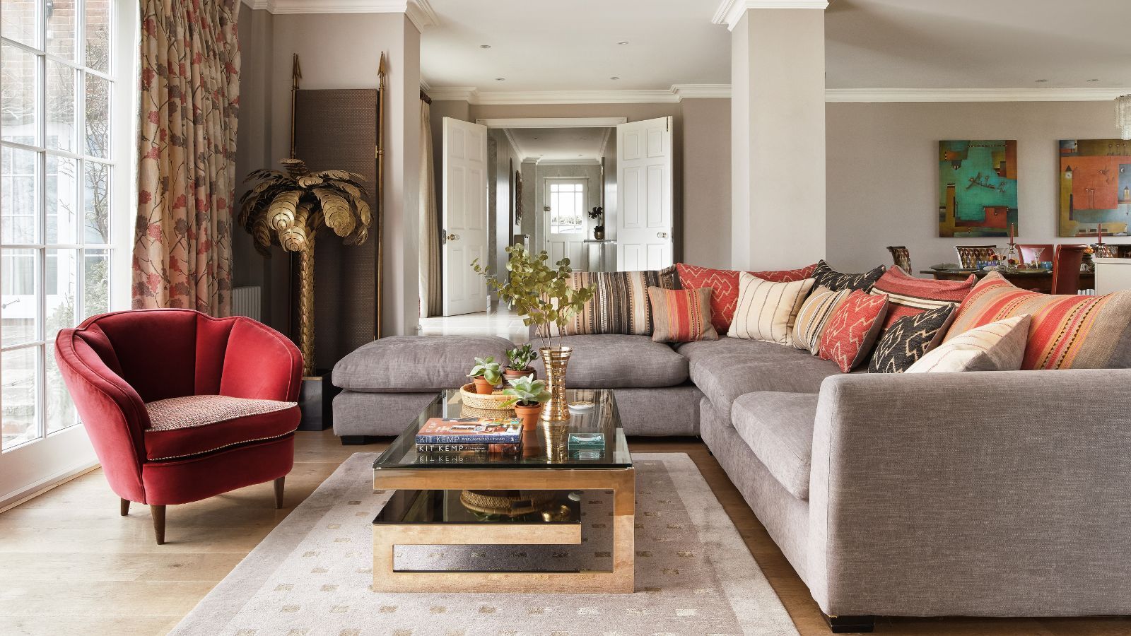 A modern living room with grey L-shaped couch, cream rug, wooden and glass coffee table, red velvet chair and gold statement palm tree in the corner.