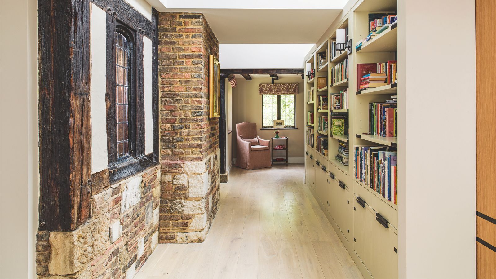 A library hallway space with bookshelves down one side, and an exposed brick wall on the other side with a Tudor-style window. At the end of the corridor is a pink upholstered chair under a window.