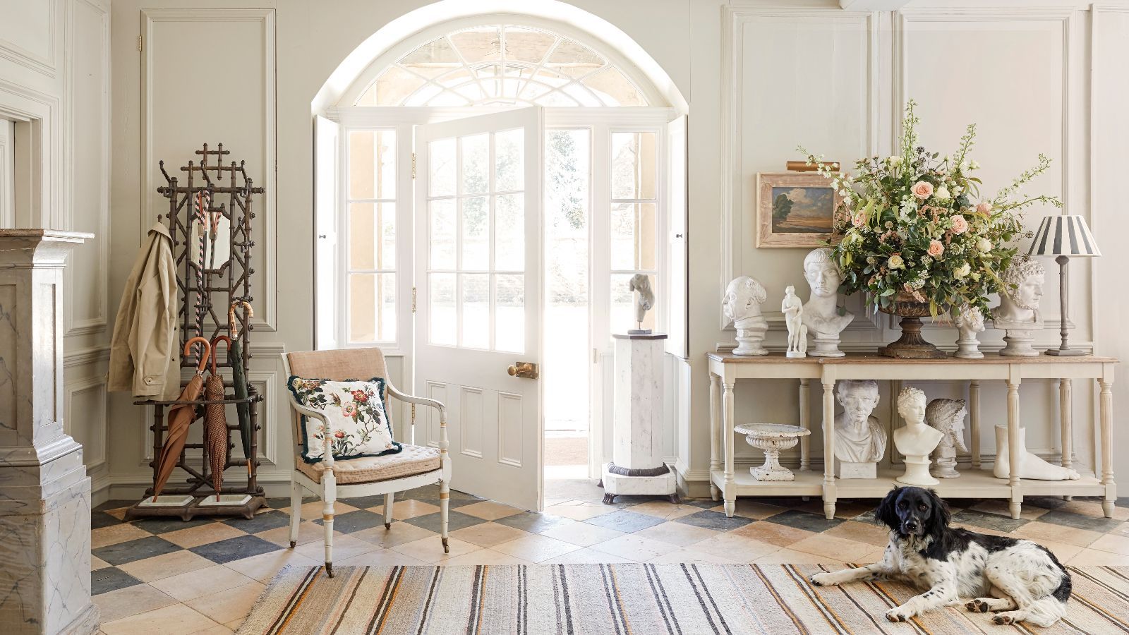 Bright and airy entryway with a striped rug on tiled patterned floor, a black and white spaniel, open glass-panelled white door, wooden umbrella stand and wooden sideboard covered in busts and flowers. The walls are white and panelled.