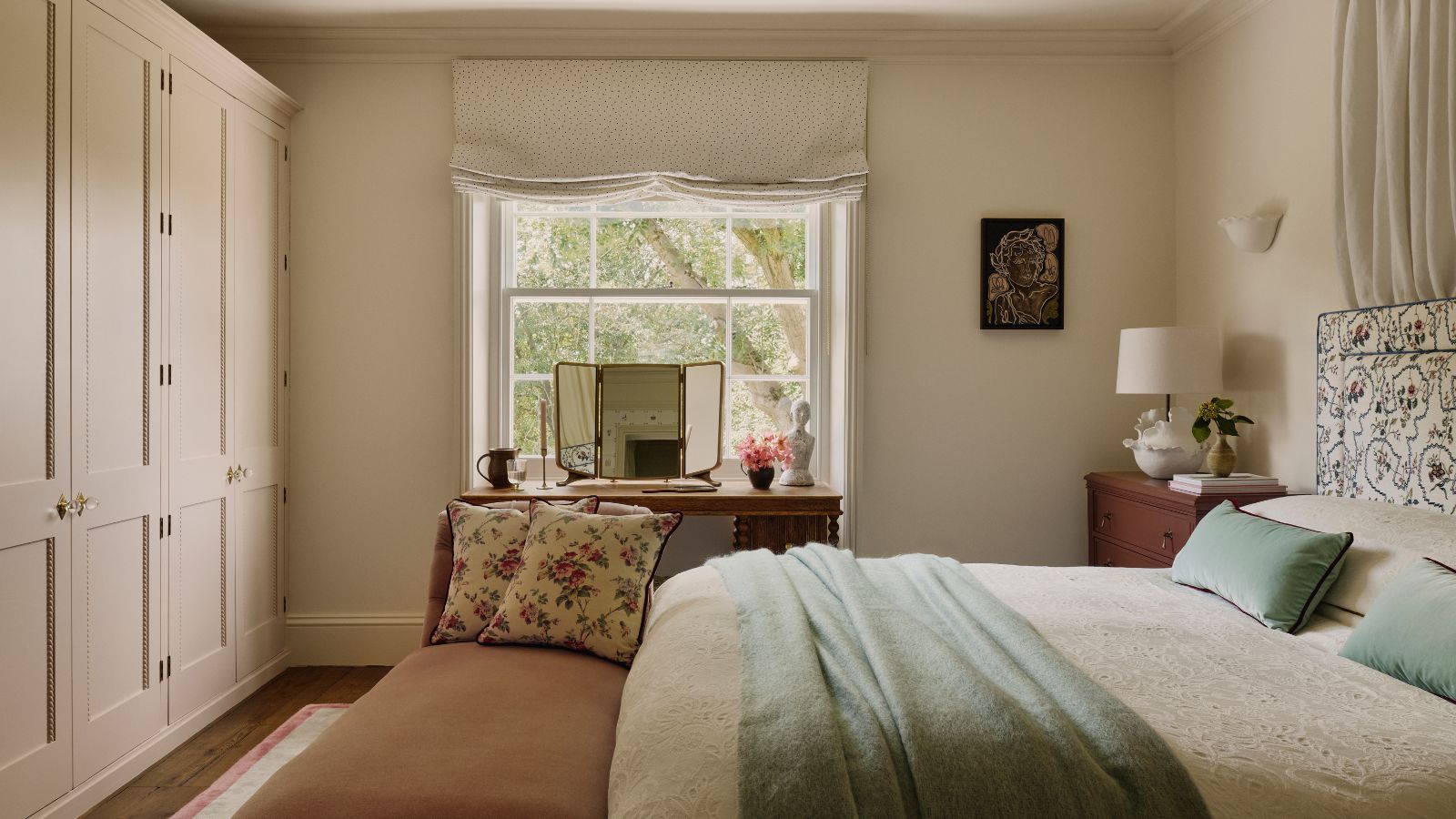 A traditional country bedroom, with built-in closets along one wall, and a bed opposite. On the far wall, a wooden dressing table is against a framed window.