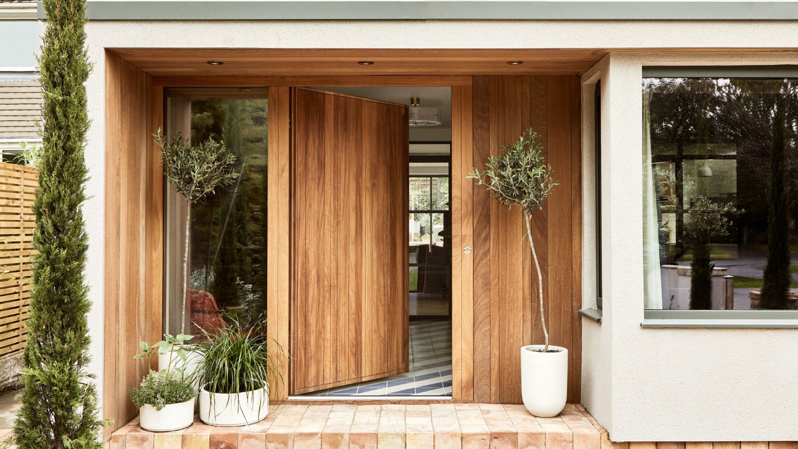 A wooden oak front door opening into a home. Plants in white pots on wither side of the door, red brick porch flooring.