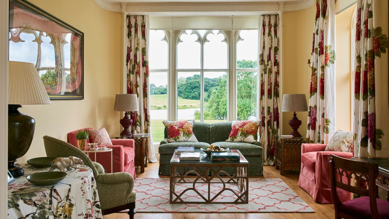 Living room with wooden floor and gothic window with views to the garden. Red and green upholstered chairs and sofa and floral curtains and cushions.