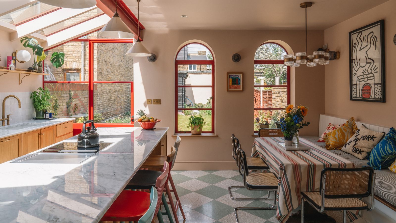A peach colored kitchen with a green and white tiled floor, large marble island, red accents on the windows, and wooden cabinetry. To the right is a large dining table with striped tablecloth, rattan chairs and bench seating, with assorted cushions and a Keith Haring artwork above.