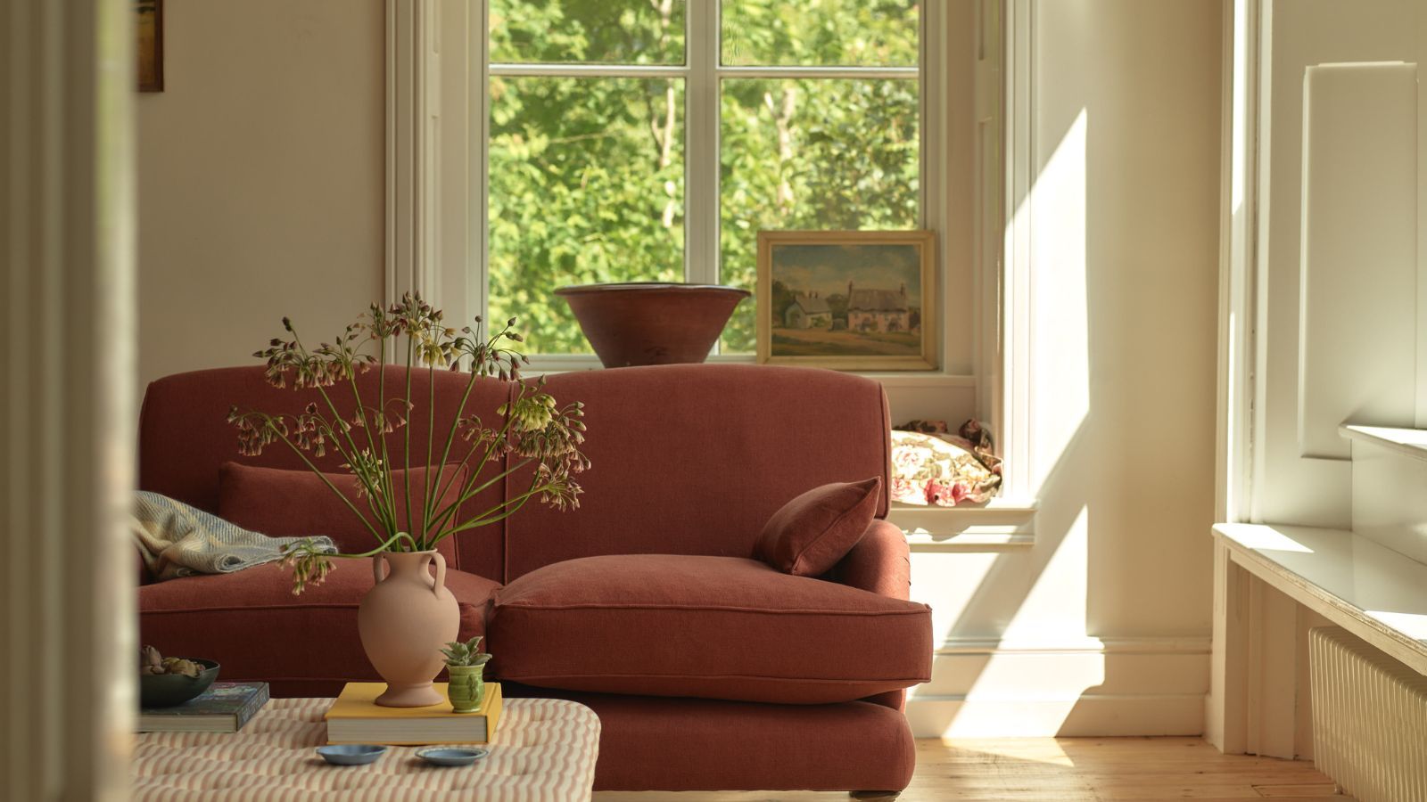A red traditional sofa in a white living room with large windows. Behind an upholstered ottoman table with a vase of large flowers on it.