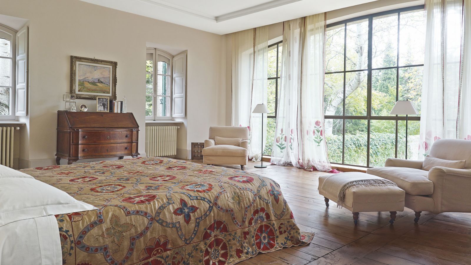 A bright bedroom with large windows with sheer curtains, brown wooden floors, a large bed with a patterned quilt, matching cream upholstered chairs, and a wooden chest of drawers against the opposite wall.