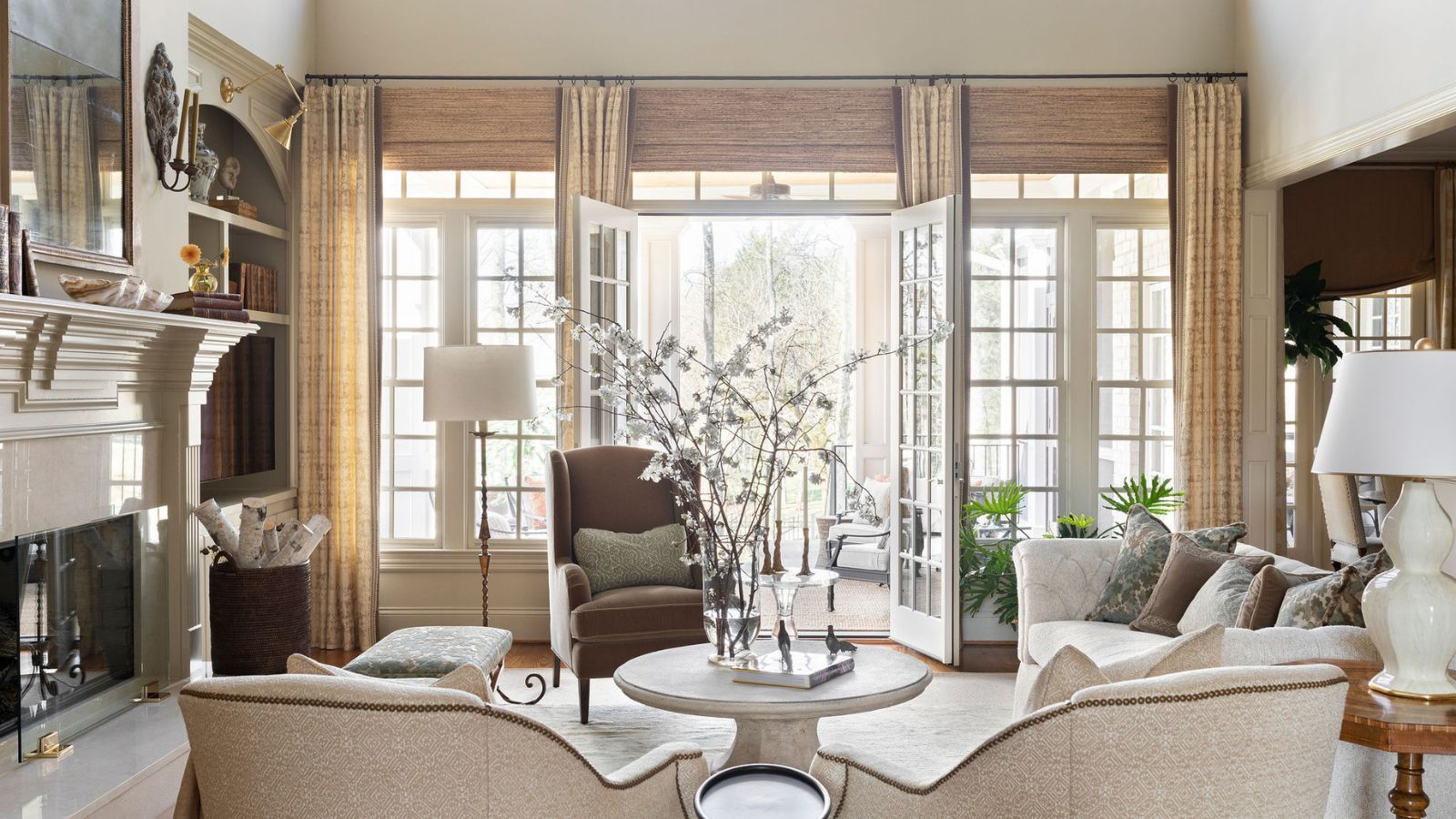 Living room decorated in neutral colours, large fireplace, beige linen sofa, armchairs, coffee table with foliage vase and large french doors