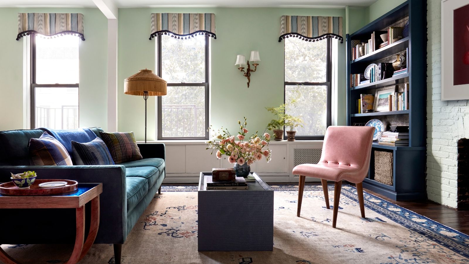 Colorful living room with coffee table, large blue sofa and pink armchair on patterned rug, in front of three windows with open blinds. A large blue bookshelf is pictured to the right.