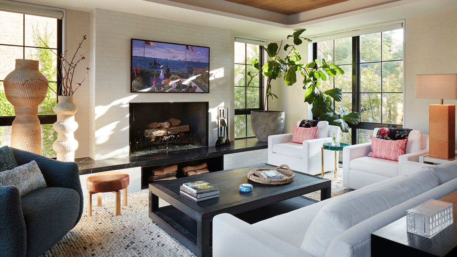 Bright living room with white brick walls, paneled windows revealing outdoor area with trees, white sofas, dark coffee table, thick rug, black armchair, TV with seaside image, curved ornaments, and a large, potted plant.
