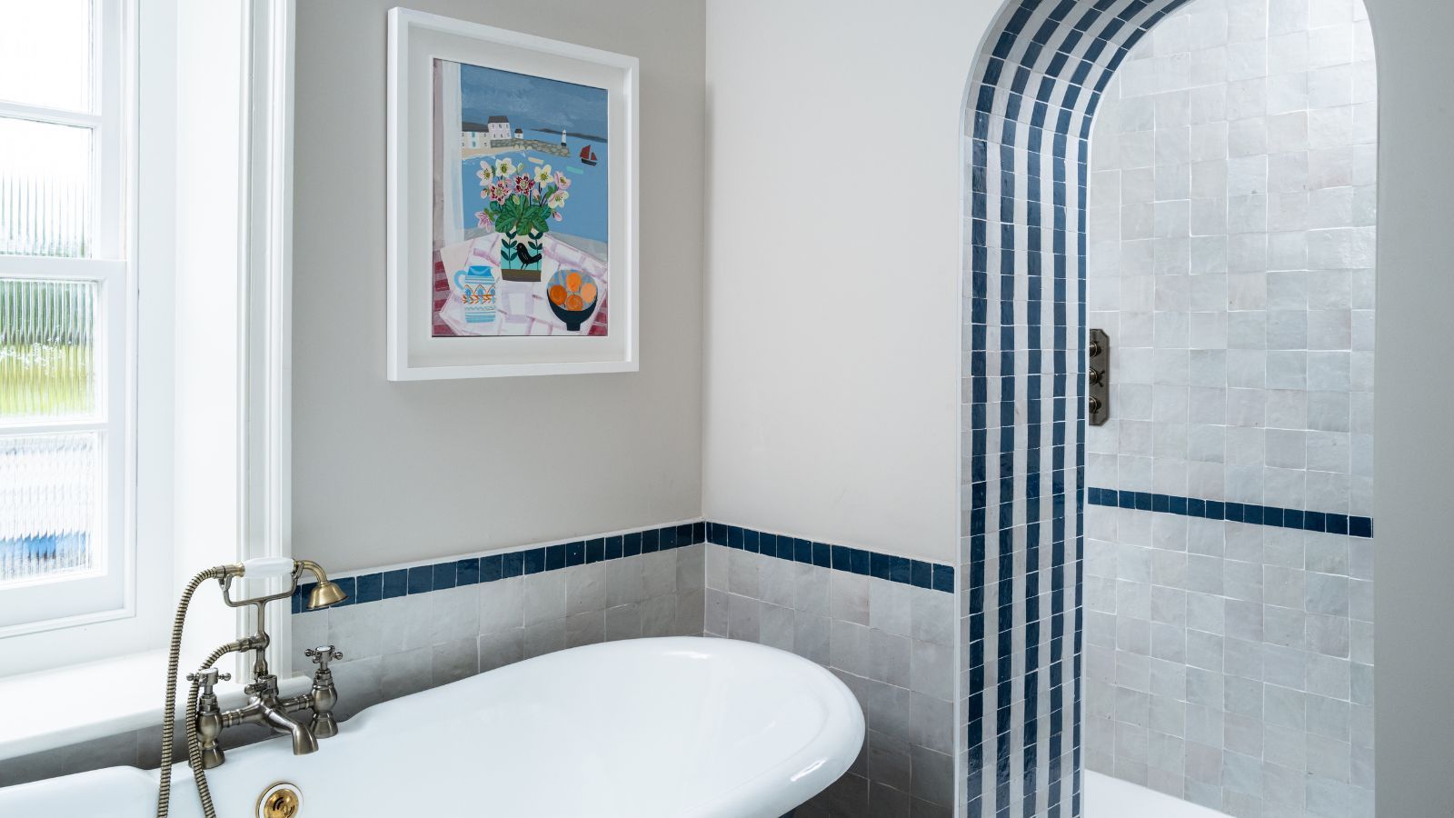 A blue and white tiled bathroom with large bathtub with gold faucets, colorful artwork, and tiled archway to a shower behind