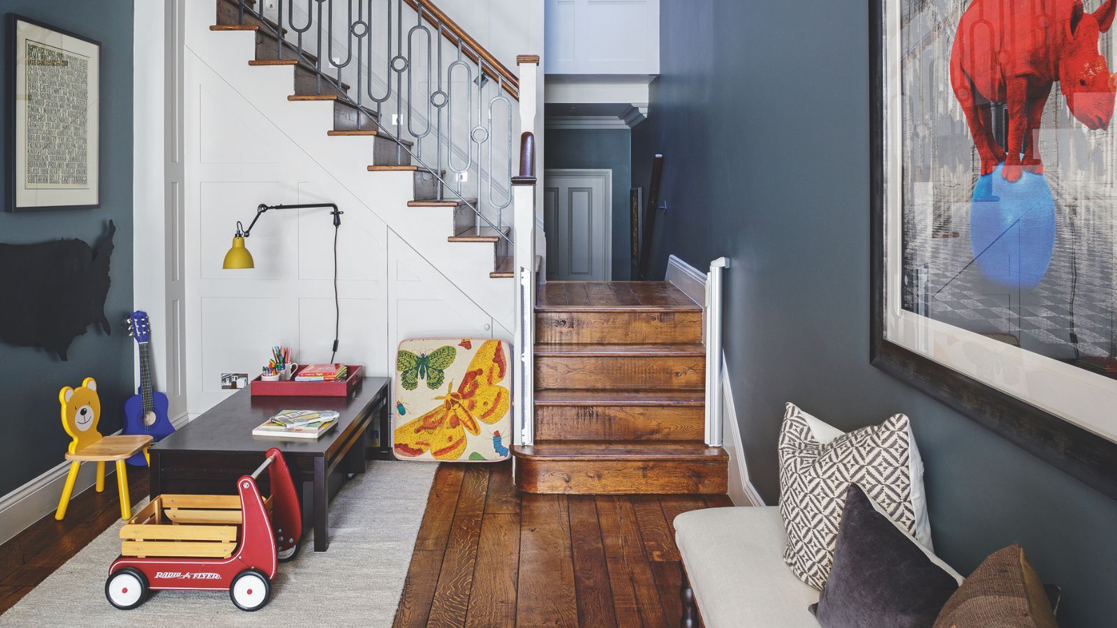 A space with dark blue walls, a white staircase, and dark wood flooring, with a children's playroom area to the left with toys and a low, dark-wood desk on a white rug. To the right is an upholstered bench with mismatched cushions, and a large artwork of a red rhino standing on a blue ball above. The staircase is seen in the background.