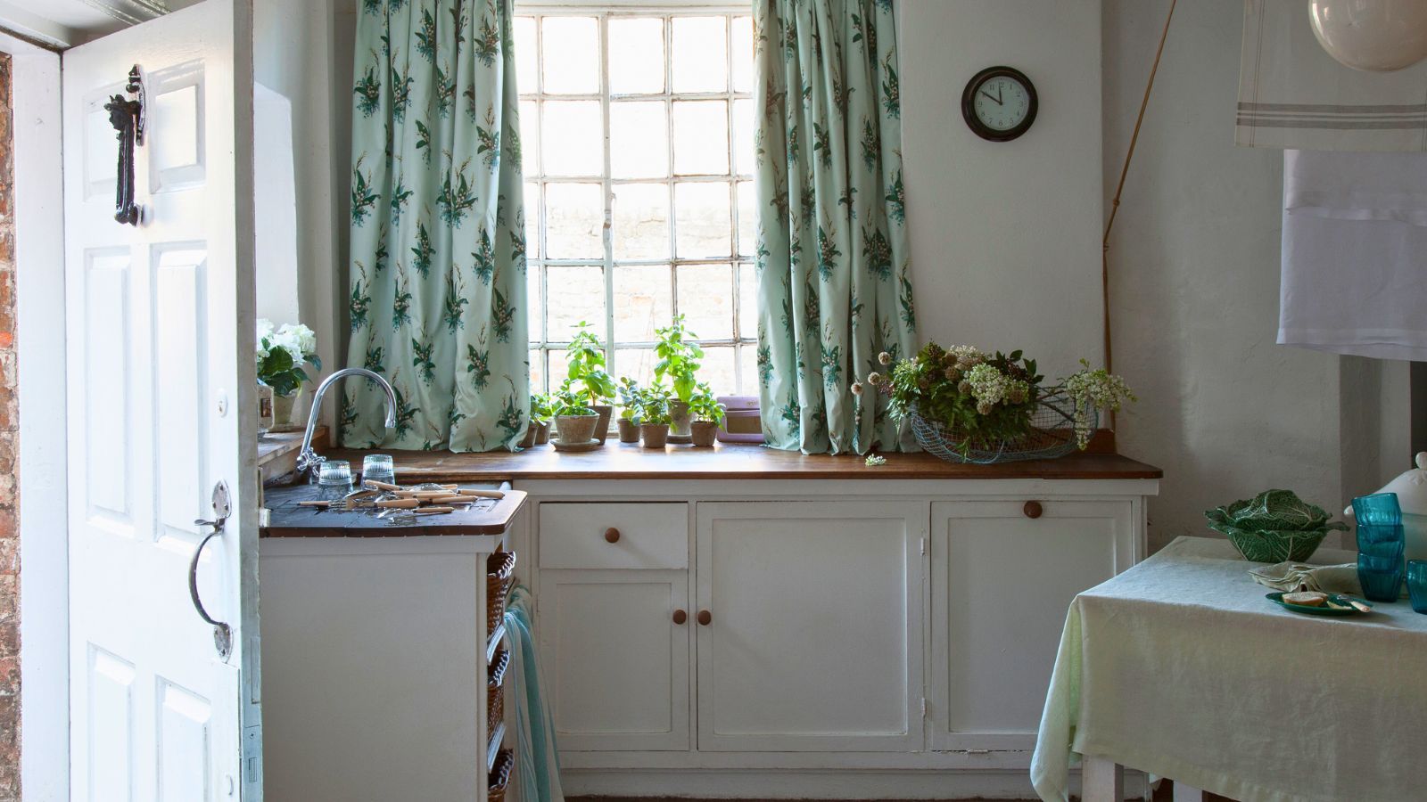 A white vintage style kitchen with wooden countertops, green curtains, white walls, a black clock and dining table covered in a green tablecloth. To the left is an open white front door.