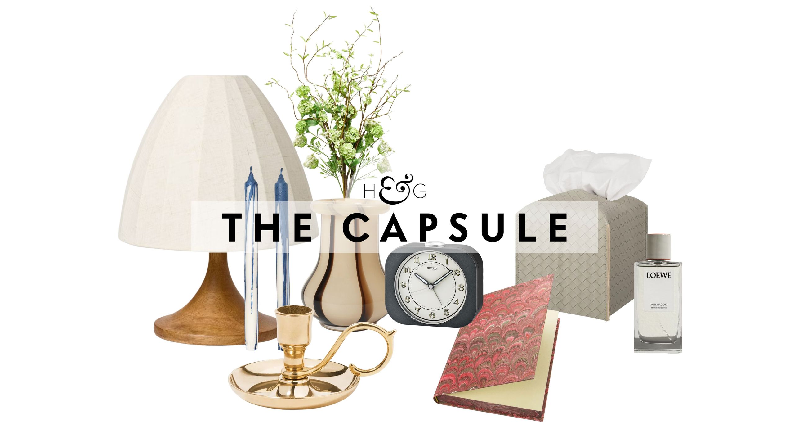 Bedside table decor pictured against a white background, including a table lamp, taper candle duo, brass candlestick, striped vase, retro-style alarm clock, patterned notebook, woven leather tissue cover, and Loewe Home spray