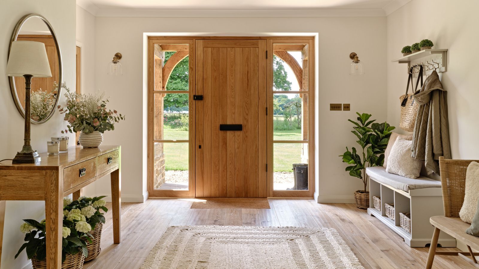 A large naturally lit entryway with a wooden front door framed by large windows. Light wooden plank flooring with a neutral boho rug. A console table on the left and coat storage on the right.