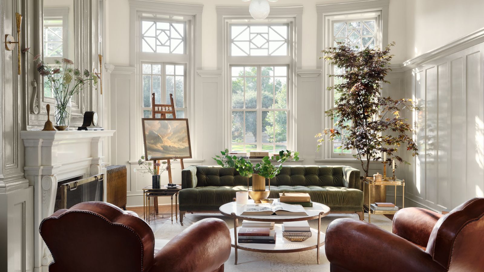 Bright and airy living room with white panelling, large windows and fireplace, leather armchairs and green velvet sofa