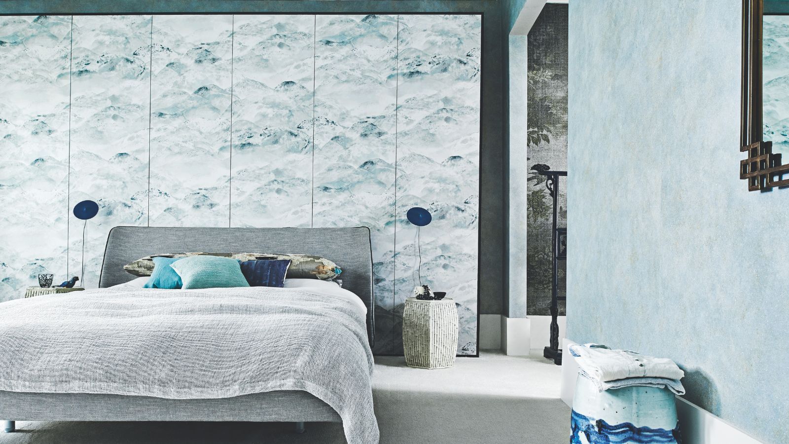 A blue bedroom with a grey double bed, statement wallpaper wall, two matching blue lamps and a blue patterned stool.