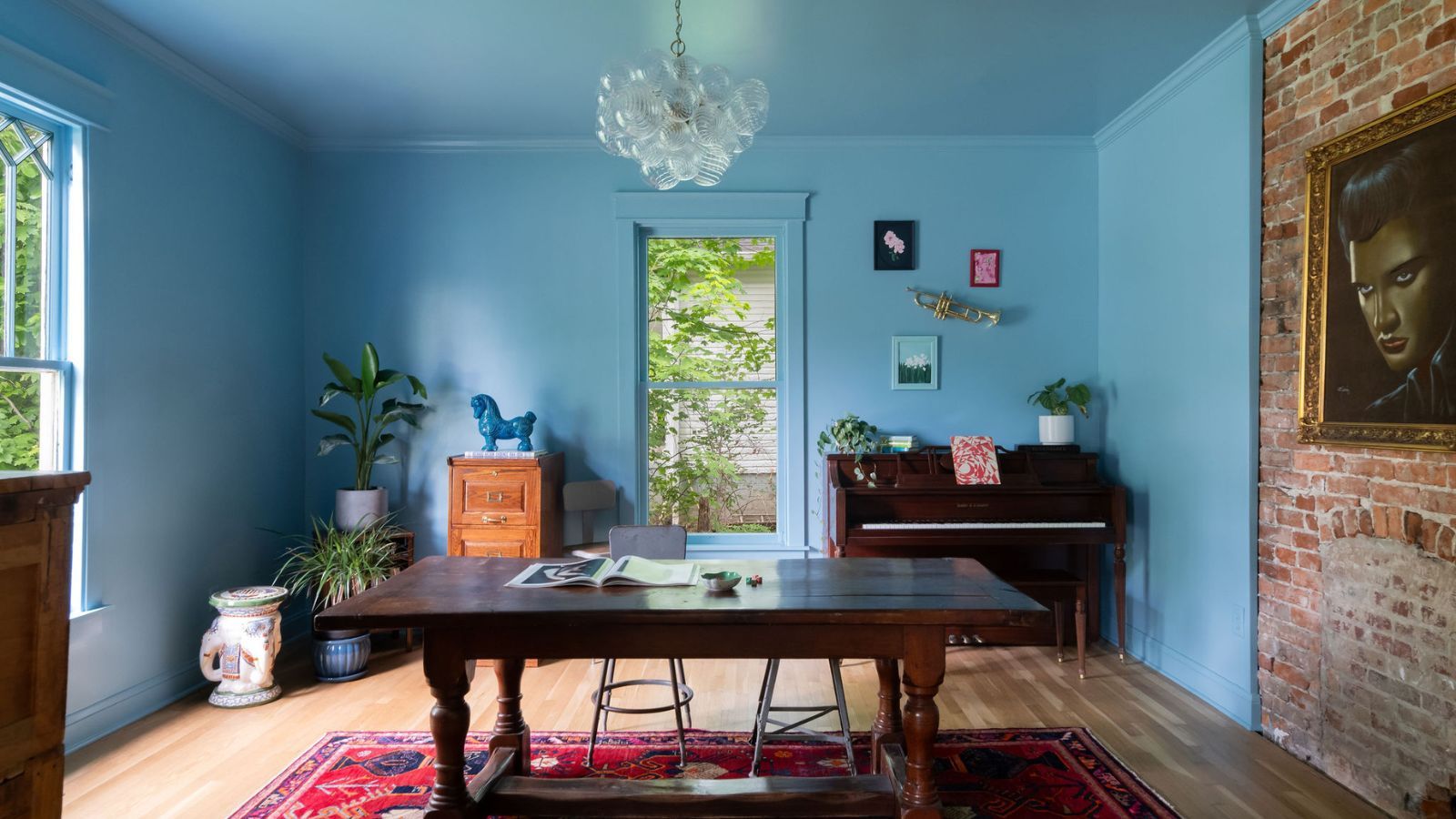 A parlor painted blue with a large wooden desk, a red rug, and a piano in the corner.