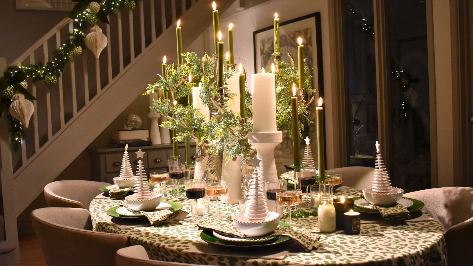 Pretty Christmas tablescape in compact open plan home with staircase in background. The table has a green and cream theme, with large church candles and ivy
