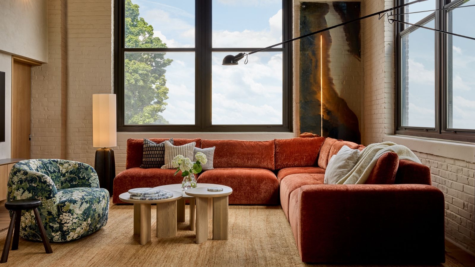 Colorful living room with orange velvet sofa, green patterned armchair, modular coffee table, and large, bright windows. Exposed brick walls and modern lighting are pictured around the furniture.