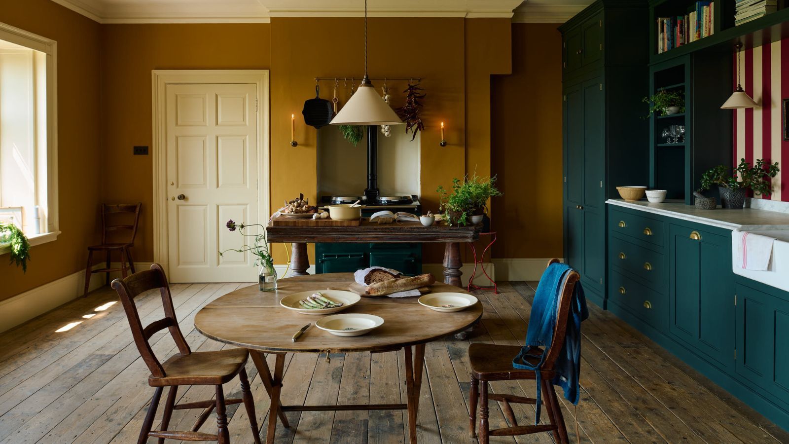 Mustard yellow and dark blue kitchen diner with hard floors and striped red and white backsplash. There are units on the right, and a window to the left, with a table in the background alongside a range cooker inset in an alcove