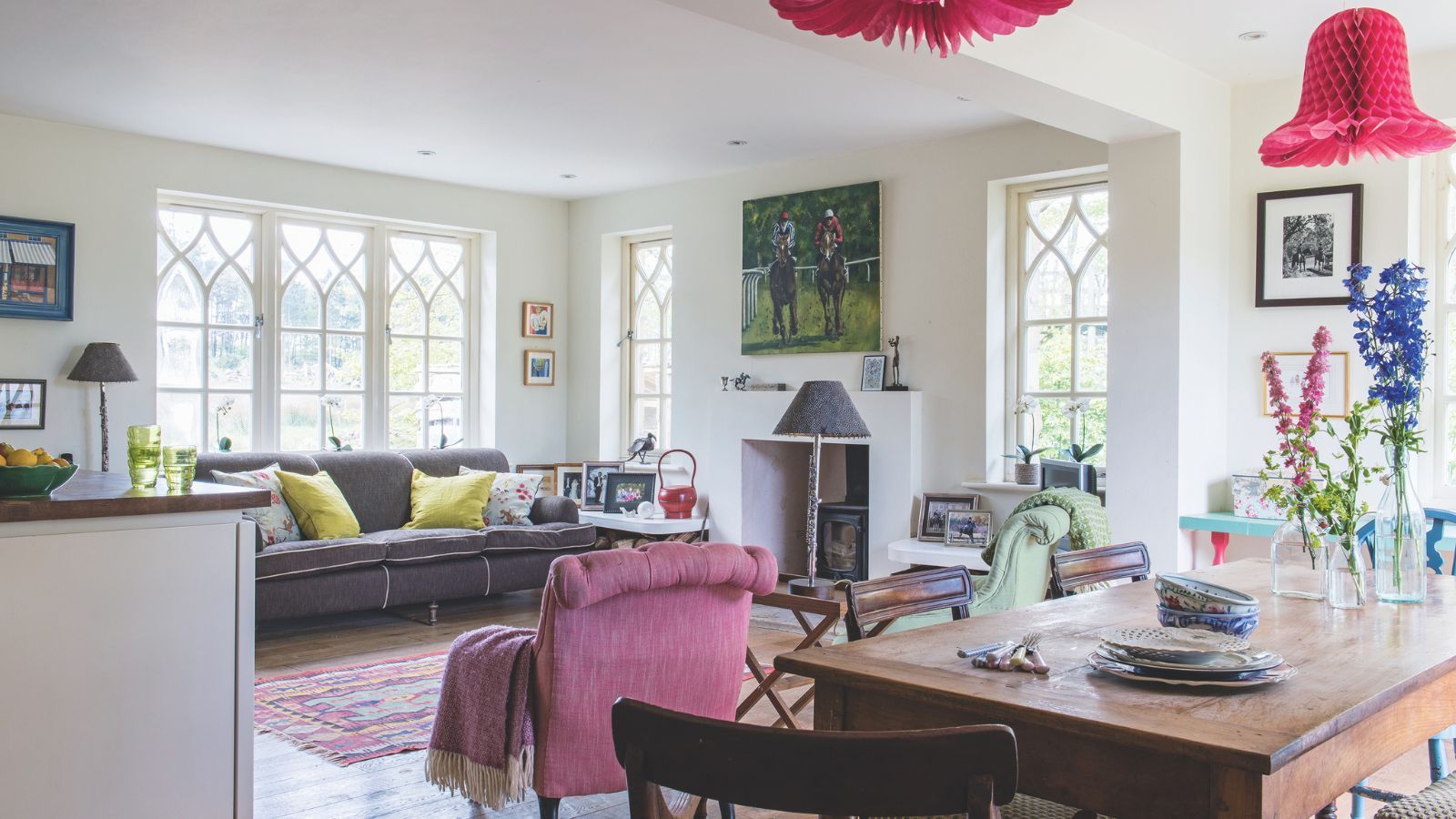A white cozy cottage style kitchen with a large wooden dining table in the foreground, white walls, surrounding latticed windows and a built-in fireplace. In the center of the room is a colorful rug and pink upholstered chair, with colorful patterns layered around the room for a comfortable maximalist look.