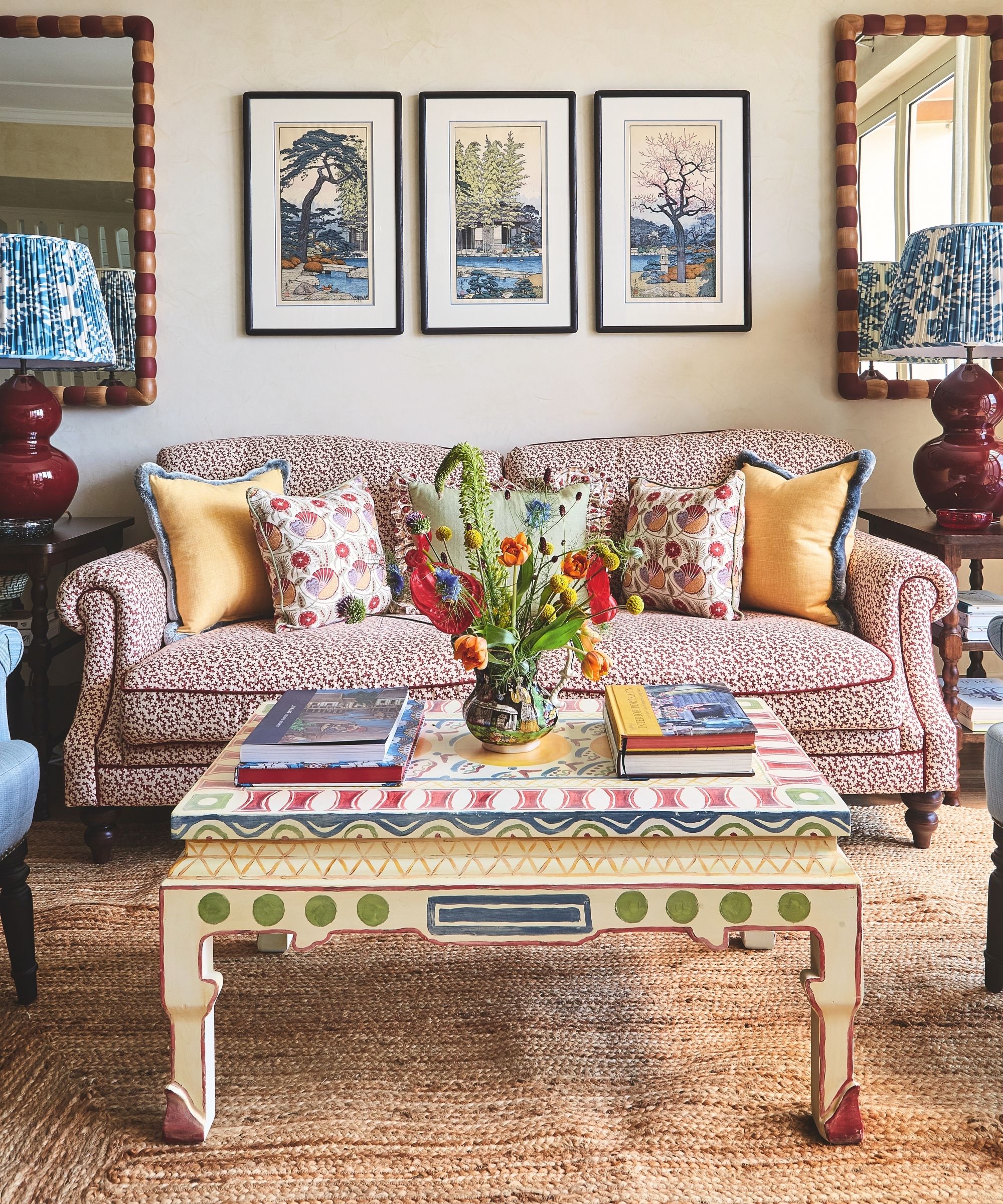 Wohnzimmer mit gemustertem Sofa und Sessel und strukturiertem Seegrasteppich und bunt bemaltem Holzcouchtisch und Kunst an den Wänden dahinter mit symmetrischen Spiegeln