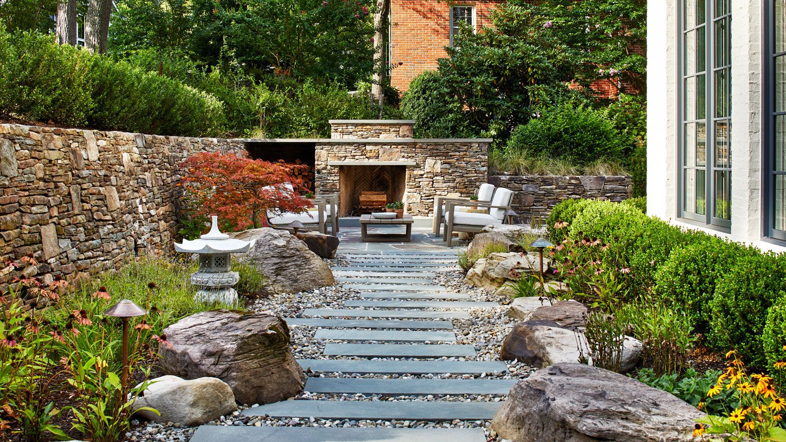 slate and gravel garden path edged with boulders and planting beds including a low hedge, and a brick wall, with an outdoor fireplace and garden furniture