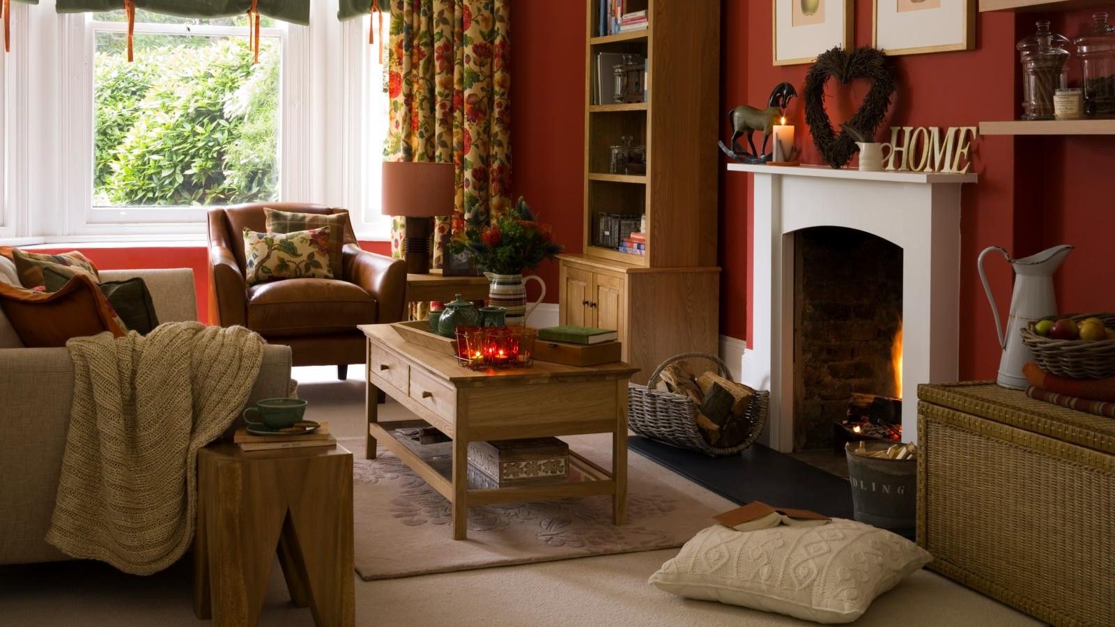 Busy living room with red walls, beige carpet and rug, wooden coffee table and two sofas. A cushion and an open book are positioned on the floor next to a burning fireplace.