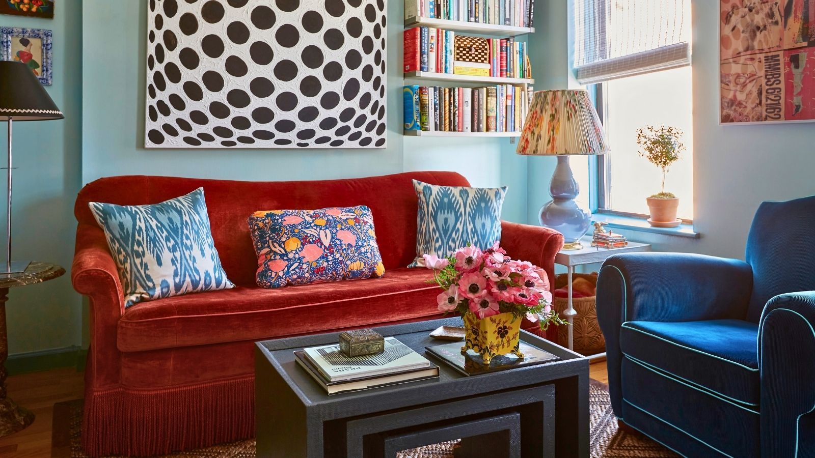 Small and cozy eclectic colorful living room with alcove shelving filled with books, orange and blue velvet sofas, and large black and white geometric artwork on the wall. There is a bright window and a coffee table with books and a vase of flowers