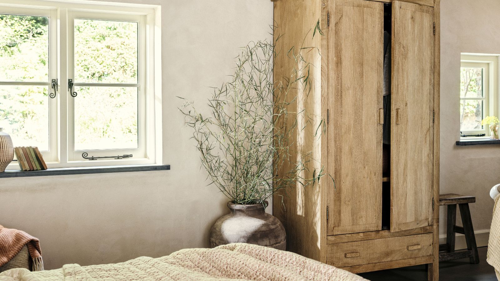 A wooden freestanding closet in a cream bedroom, with a green spindly plant in a stone pot beside it.