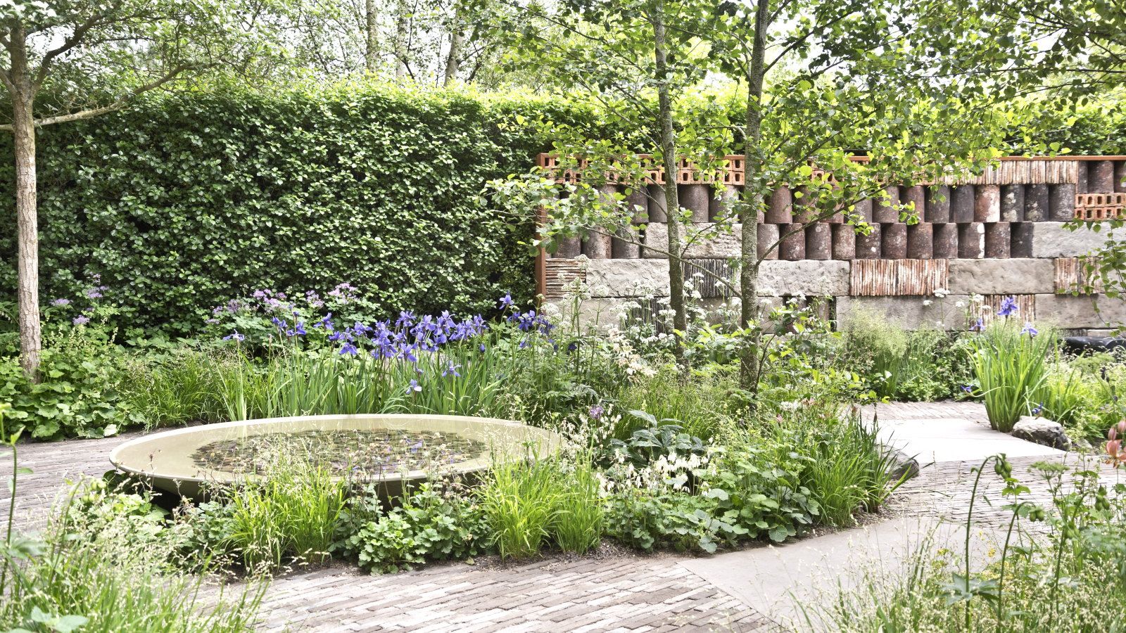A show garden complete with a brick patio, circular water feature, borders, and a large hedge at the back