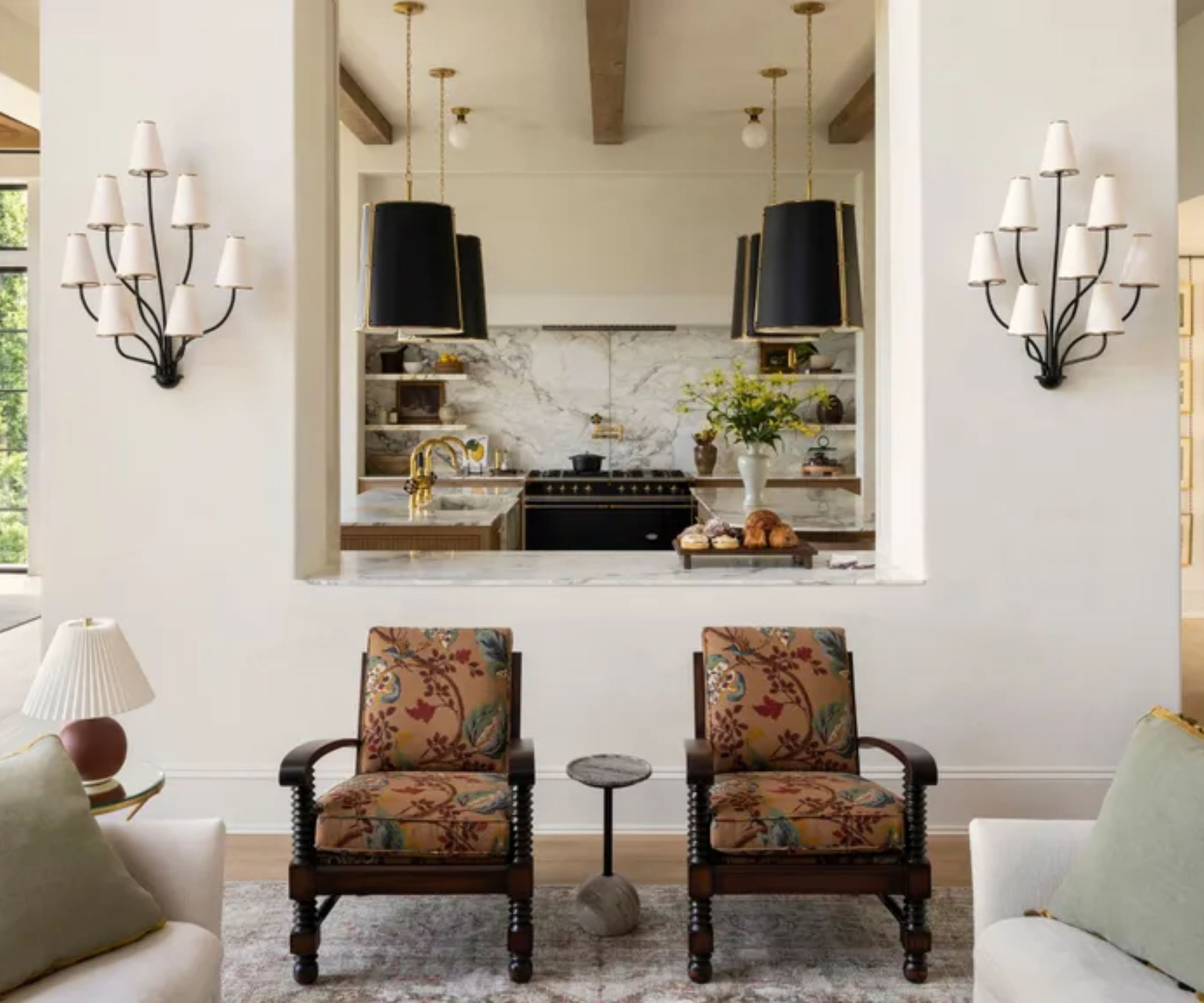 Ein helles Wohnzimmer mit zwei weißen Sofas vor einem Steinkamin. In der Ecke steht ein großer Baum und am Kamin stehen Regale mit Töpferwaren. Darüber hängt ein runder goldener Kronleuchter und hohe, schwarz gerahmte Fenster lassen natürliches Licht herein. In der Mitte steht ein Couchtisch mit Büchern und Blumen.