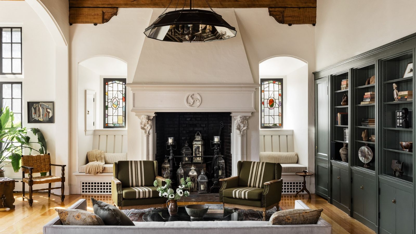 Living room with green built-in bookshelves, two striped green armchairs, an ornate wall with stained glass windows, and miscellaneous flowers and ornaments.